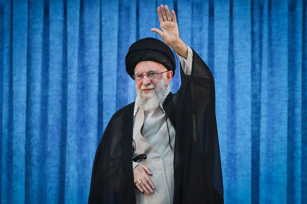 O ex-l&iacute;der supremo do Ir&atilde;, o aiatol&aacute; Ali Khamenei, de p&eacute;, com a m&atilde;o esquerda erguida, em frente a uma cortina azul, durante uma cerim&ocirc;nia no dia 4 de junho de 2025