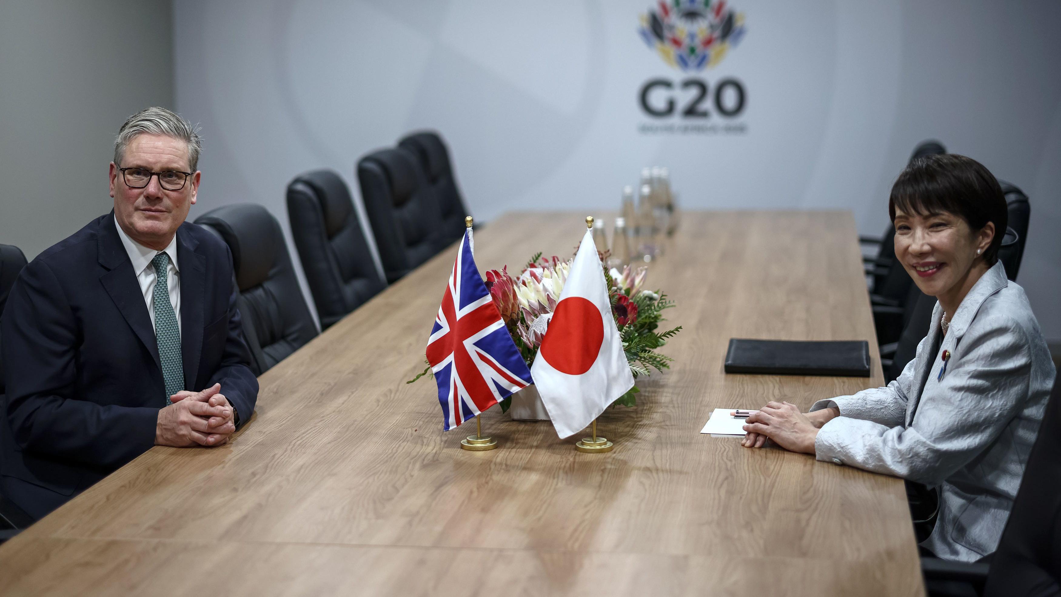 施紀賢爵士（左）與高市早苗（右）在南非約翰內斯堡G20峰會週邊會晤，兩人坐在長會議桌中央兩側，桌面中間擺放著英國與日本小型國旗（22/11/2025）