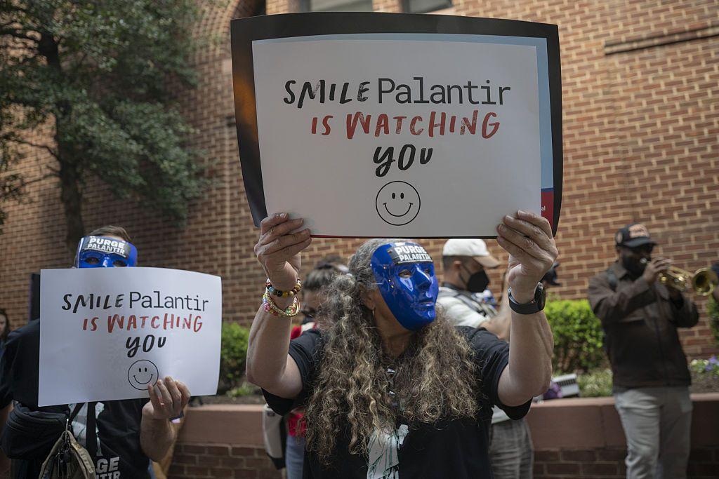 Manifestantes con máscaras azules sostienen letreros que leen "Sonríe, Palantir te está mirando".