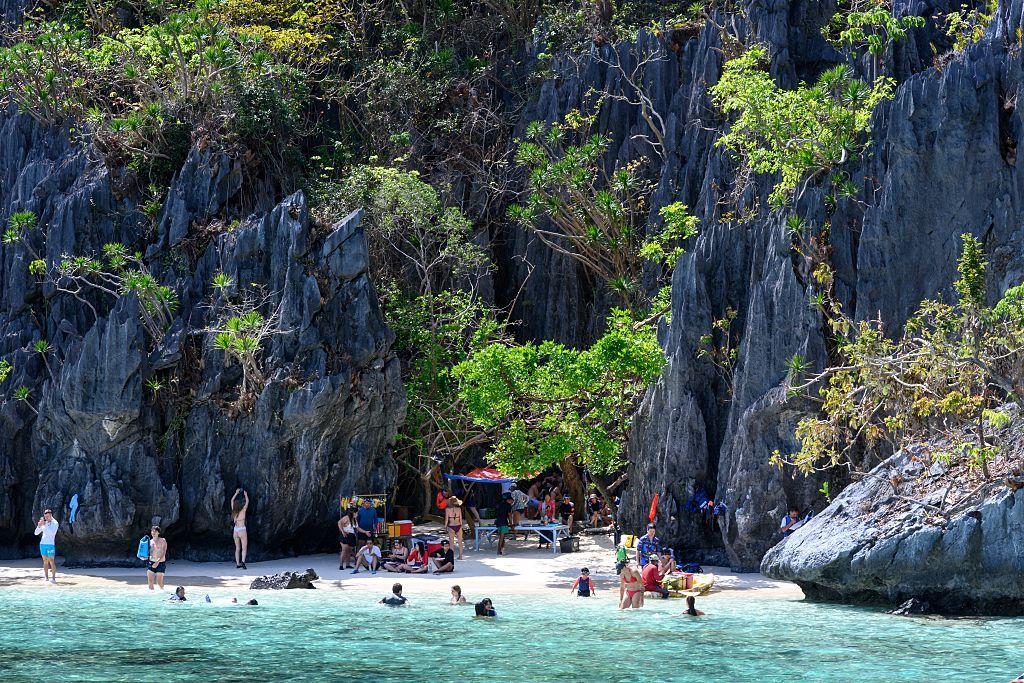 Pequeña playa entre rocas empinadas y algunos árboles, con varias personas nadando y en la arena, así como una mesa y venta de algo que no se alcanza a ver