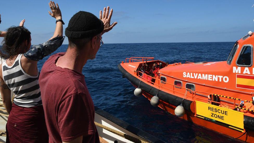 Duas pessoas acenam para um barco laranja da guarda costeira, com bandeira espanhola