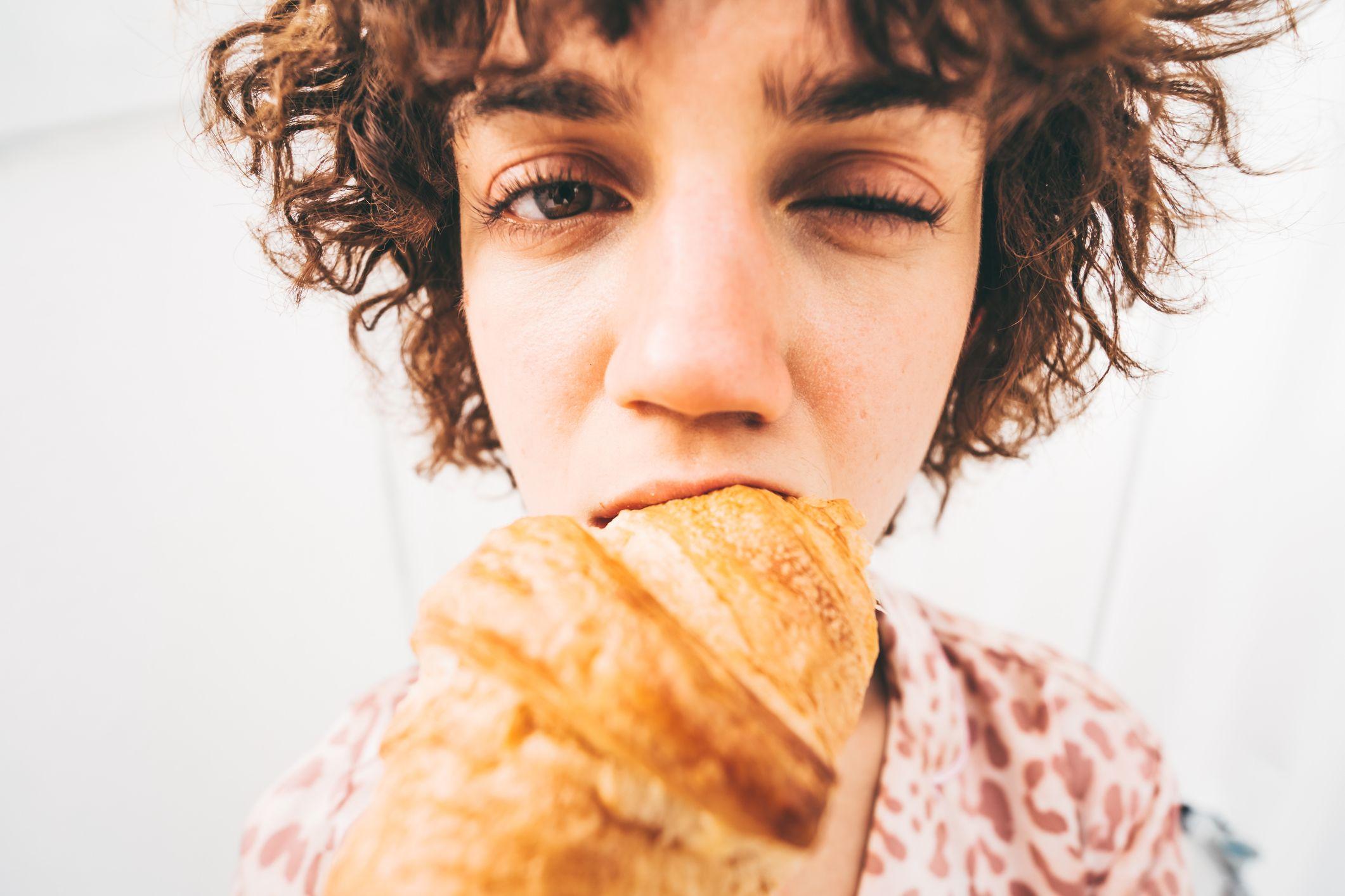 Žena jede kroasan, trepćući jednim okom dok to radi. Fotografija „riblje oko“ preuveličava njeno lice, kroasan i zalogaj