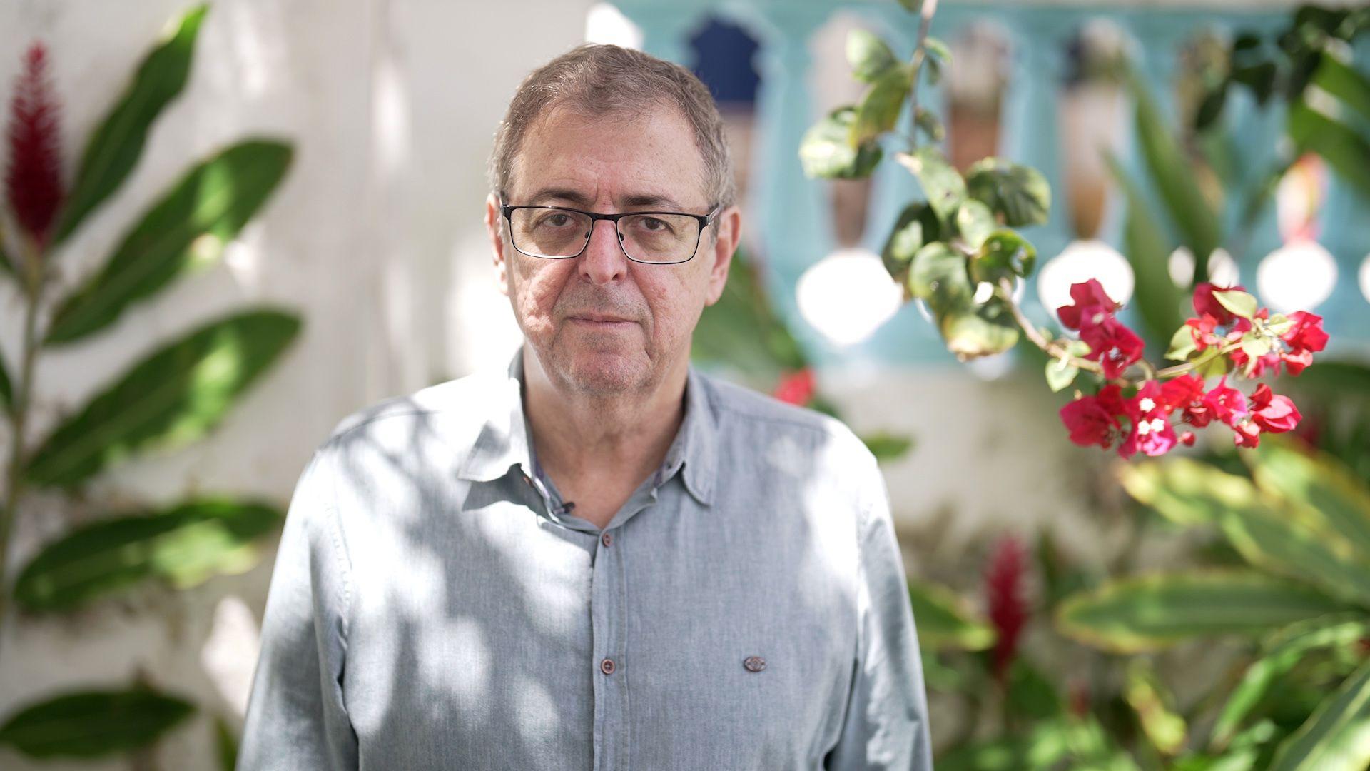 Homem branco, de óculos, com camisa cinza e flores roxas à direita e ao fundo