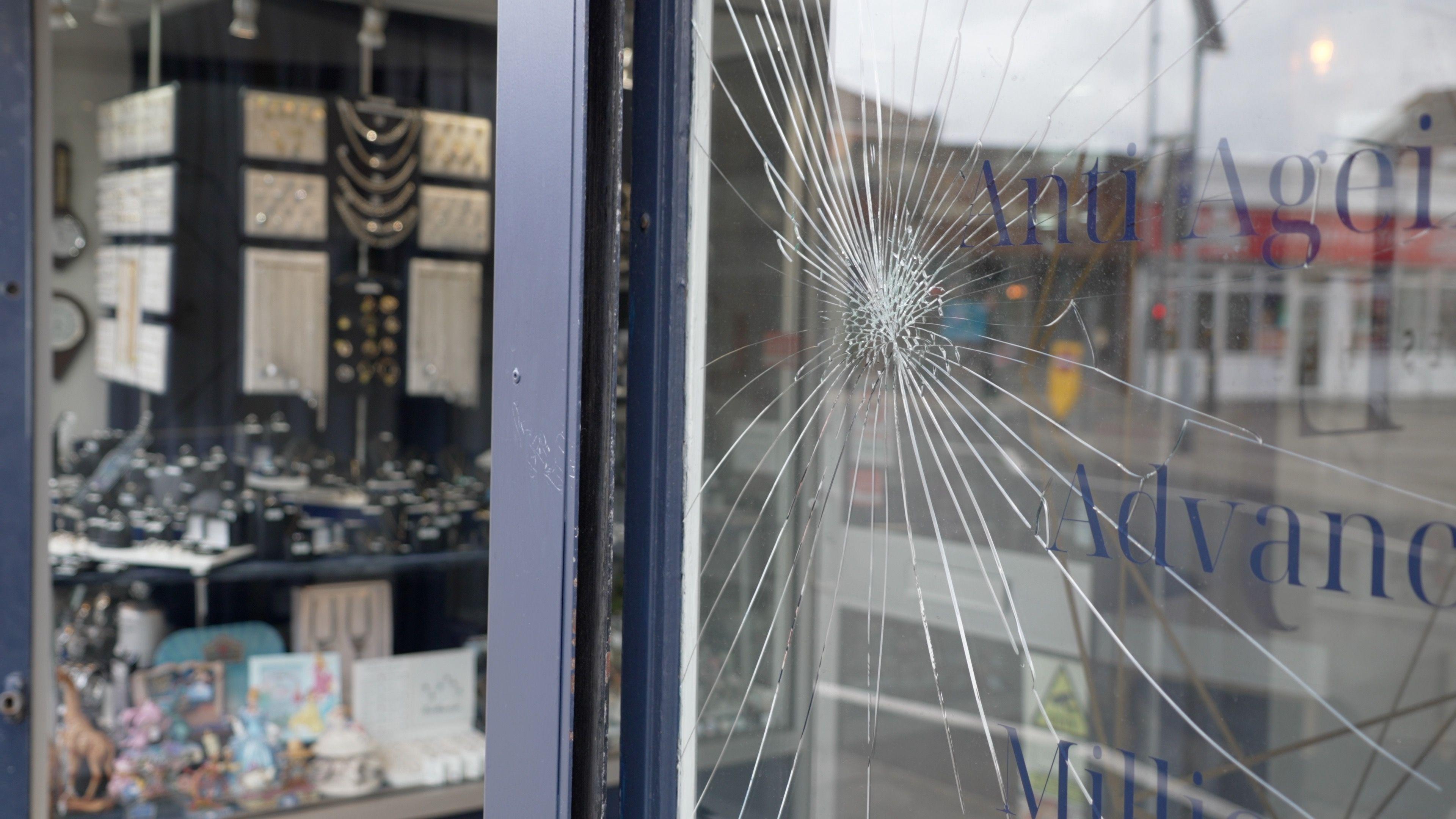 Vitrine rachada de uma loja é mostrada do lado de fora, com o reflexo da rua