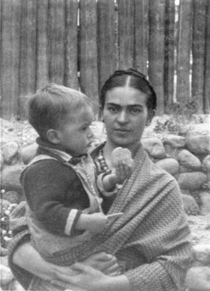 Frida mira a la cámara mientras tiene cargado al pequeño Antonio, que tiene una galleta en la mano. Atrás hay una pared de piedras y una valla de madera.

