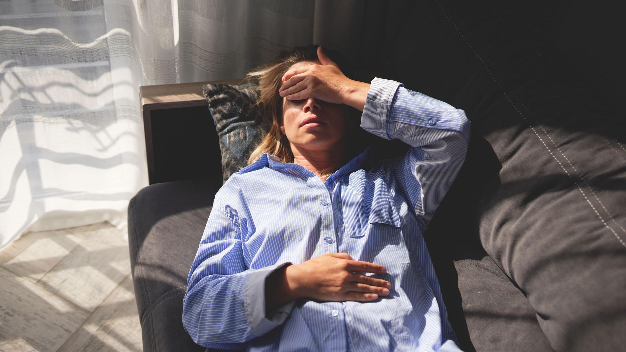 Mulher deitada em um sof&aacute;, aparentemente com dor de cabe&ccedil;a, ao lado de uma janela por onde entra forte luz do dia