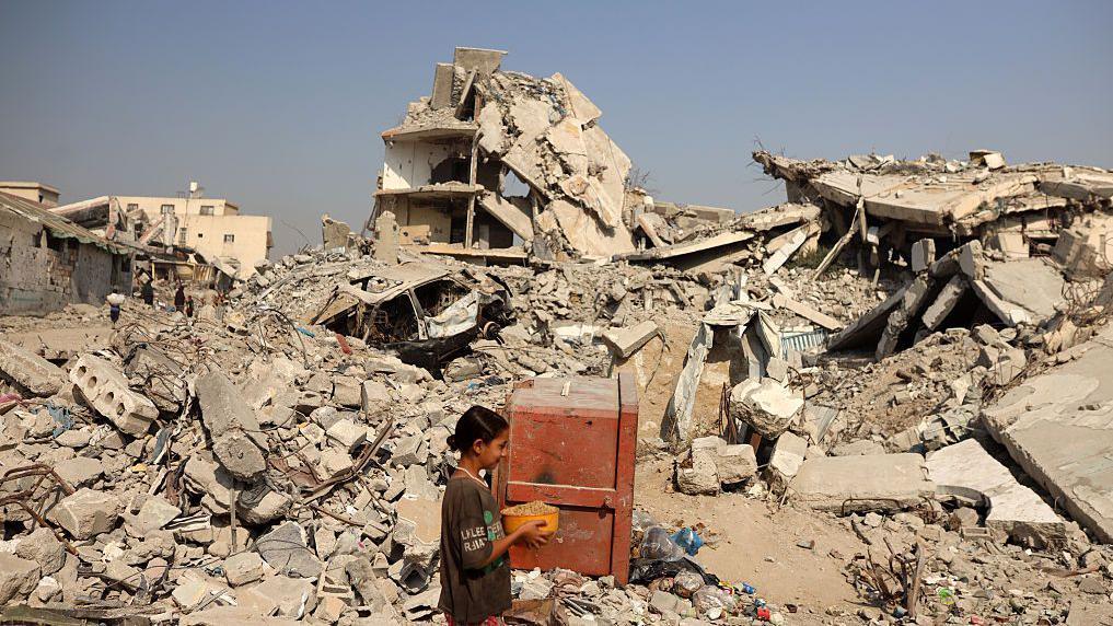 Una niña camina entre edificios destruidos con una olla entre las manos  en Ciudad de Gaza. 
