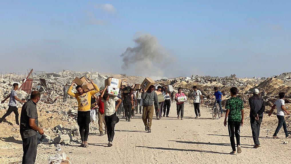People walking along a dusty road carrying boxes of aid from a GHF centre in late May as smoke rises in the background