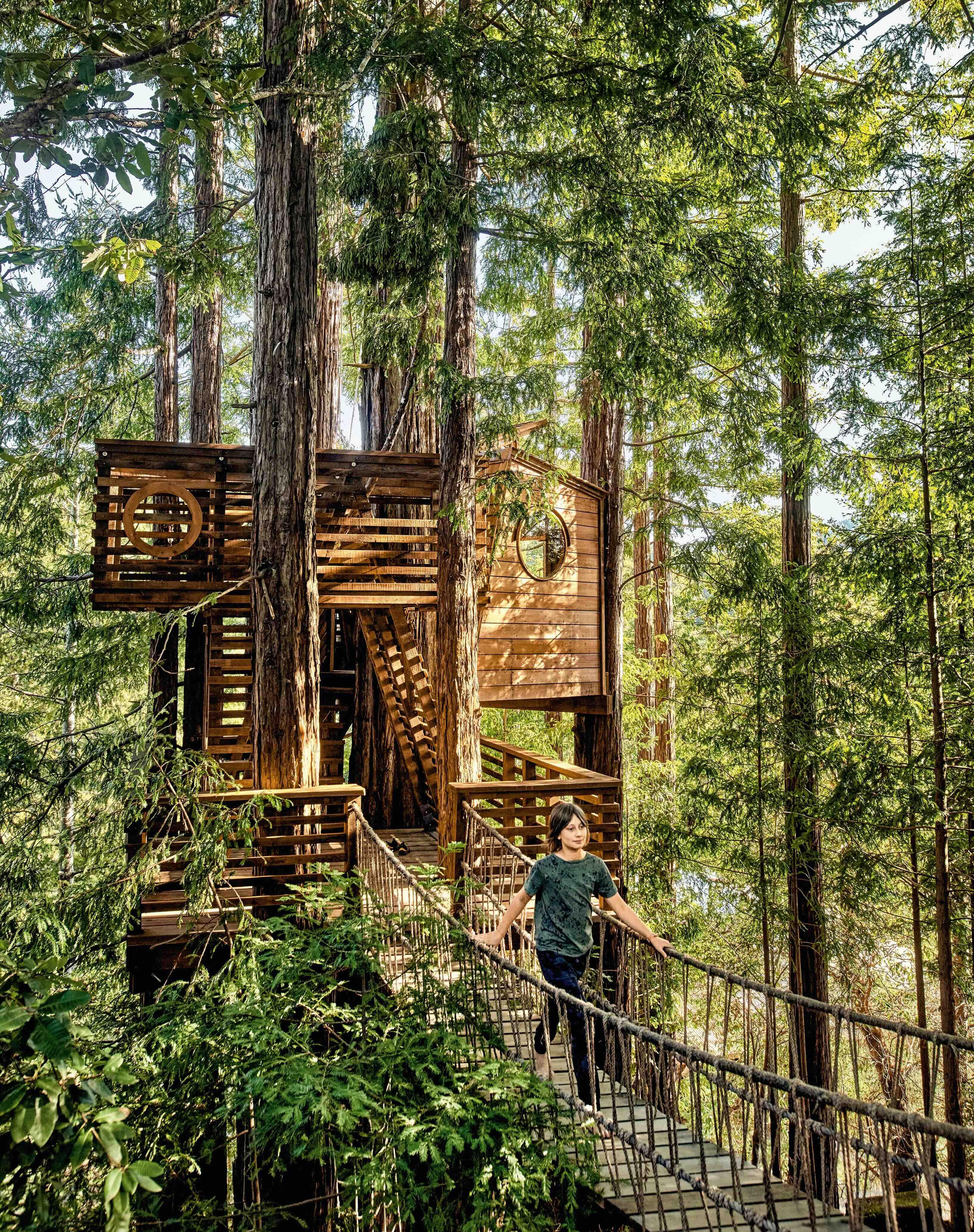 Una casa en un árbol de madera, de dos pisos, que está conectada a un puente colgante por el cual camina una mujer. 