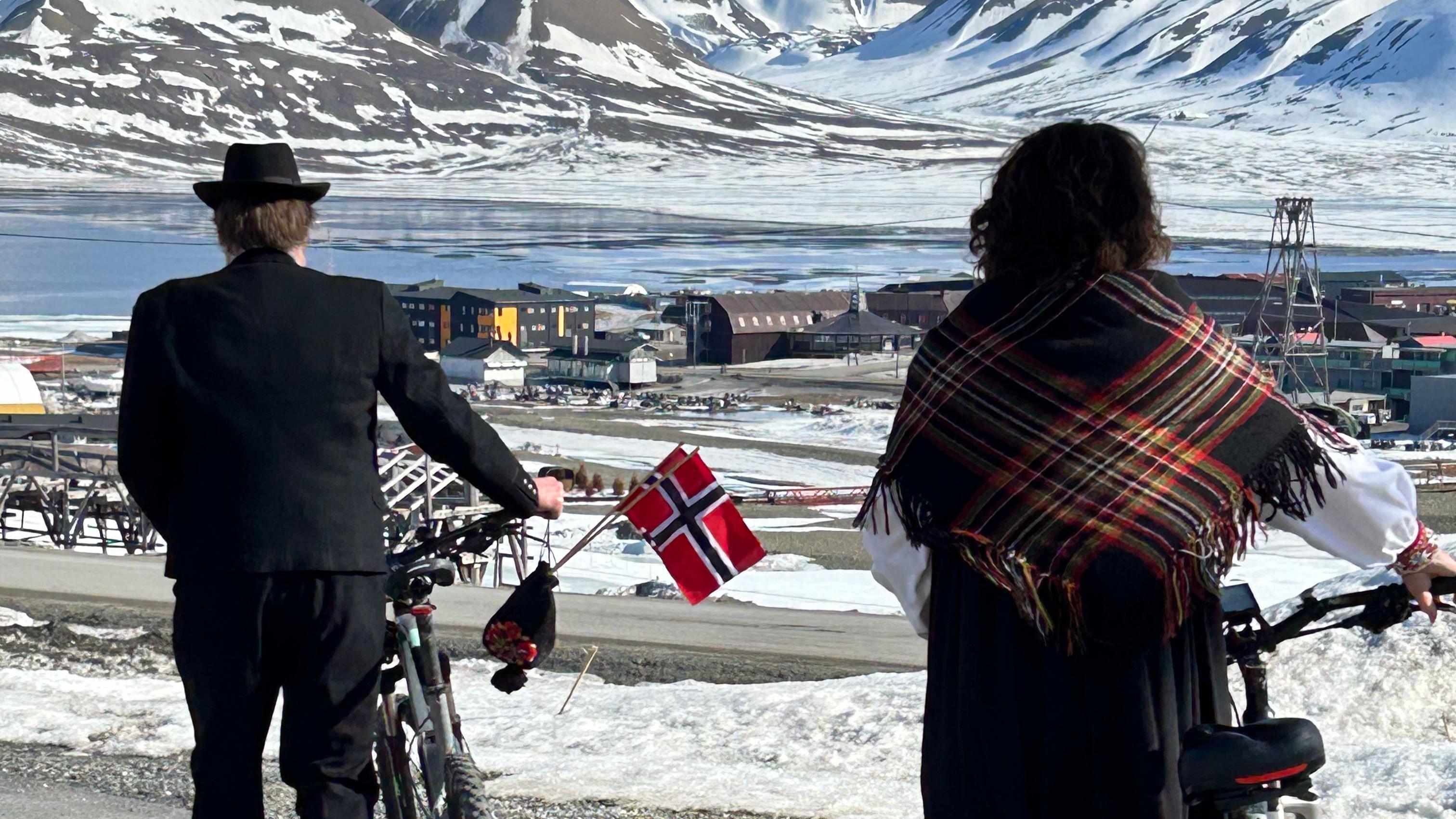Dos noruegos paseando en bicicleta en su día nacional en Svalbard, con agua y picos helados a lo lejos.