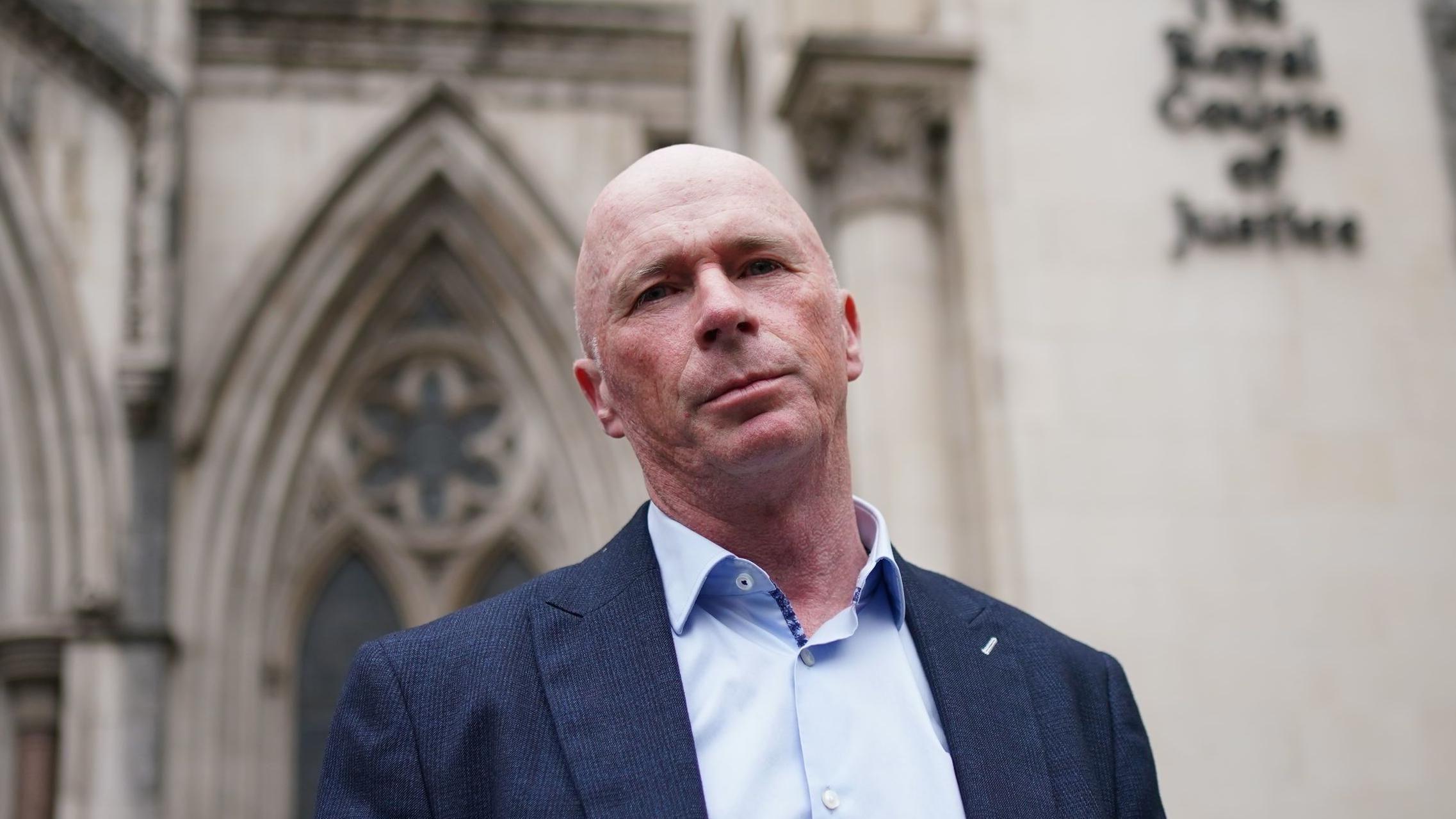 صورة لريموند كونولي وهو يقف خارج مجمع المحاكم الملكية للعدل في لندن، مرتدياً قميصاً أزرق فاتحاً وسترة زرقاء داكنة. 