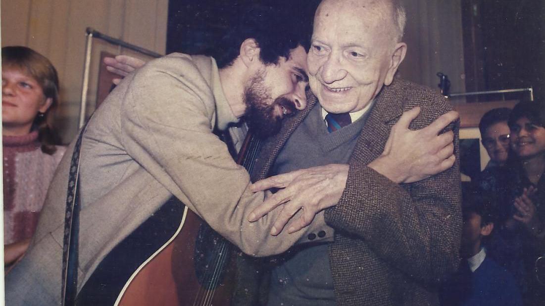 A fotografia mostra um homem jovem com um viol&atilde;o nas costas, Henrique Mann, abra&ccedil;ando um homem idoso, Mario Quintana, que sorri durante o encontro. Ao fundo, outras pessoas acompanham a cena em um ambiente interno, sugerindo um evento ou homenagem marcada por um momento de afeto e celebra&ccedil;&atilde;o.