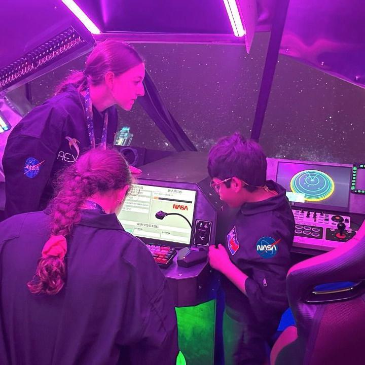 David Camacho, com duas jovens, todos vestidos com uniformes da Nasa, dentro de uma cabine espacial, observando um computador