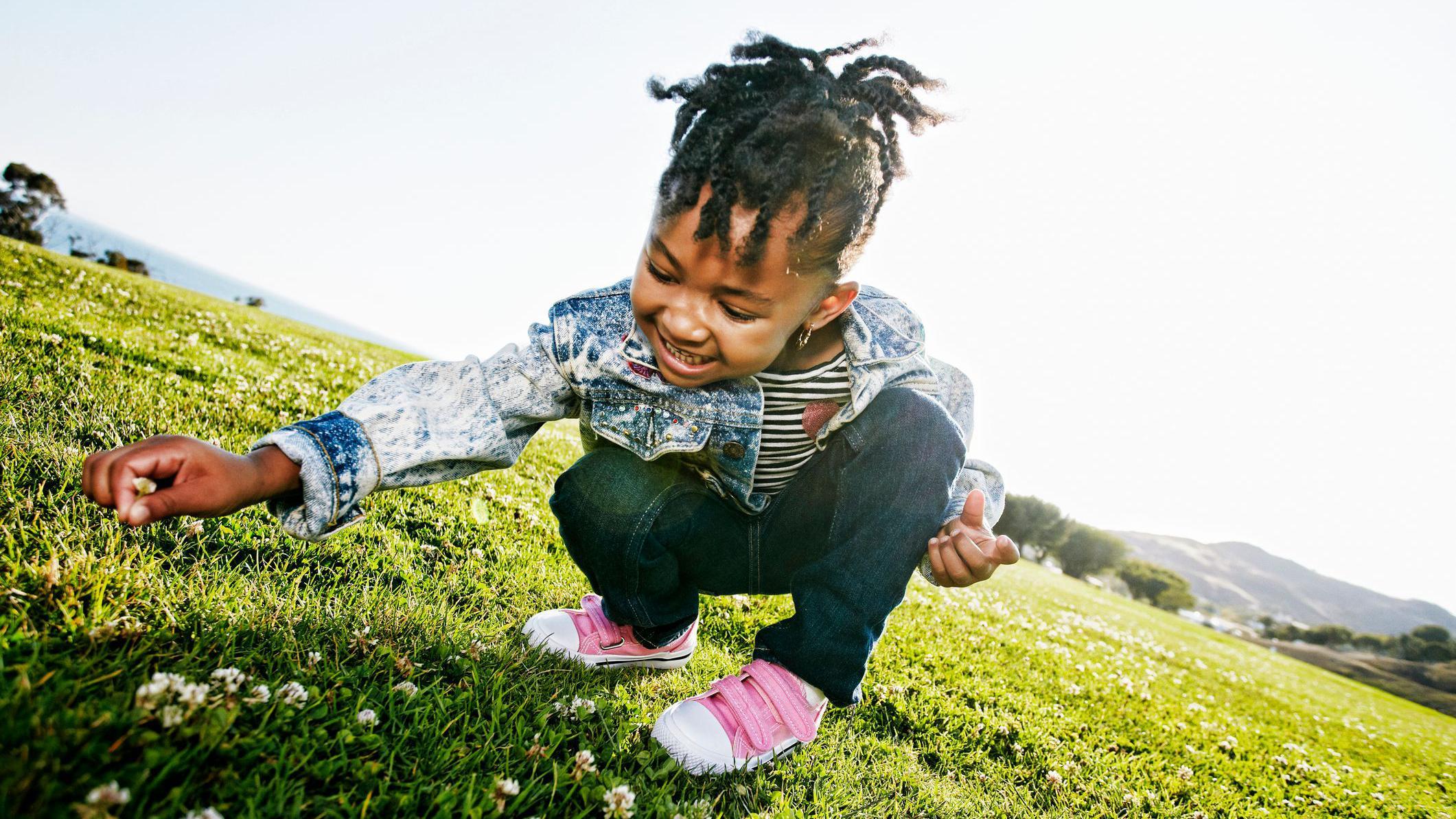 Una niña sonríe mientras está agachada en cuclillas profundas sobre un campo de césped iluminado por el sol. Está buscando pequeñas flores blancas. La niña lleva una chaqueta vaquera, pantalones oscuros y zapatos rosas. 