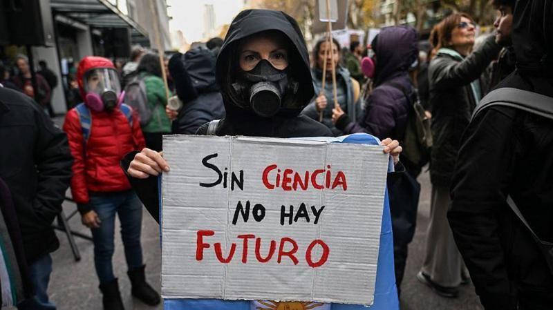 Manifestante com capuz e máscara contra gases carrega uma bandeira da Argentina e um cartaz com os dizeres 
