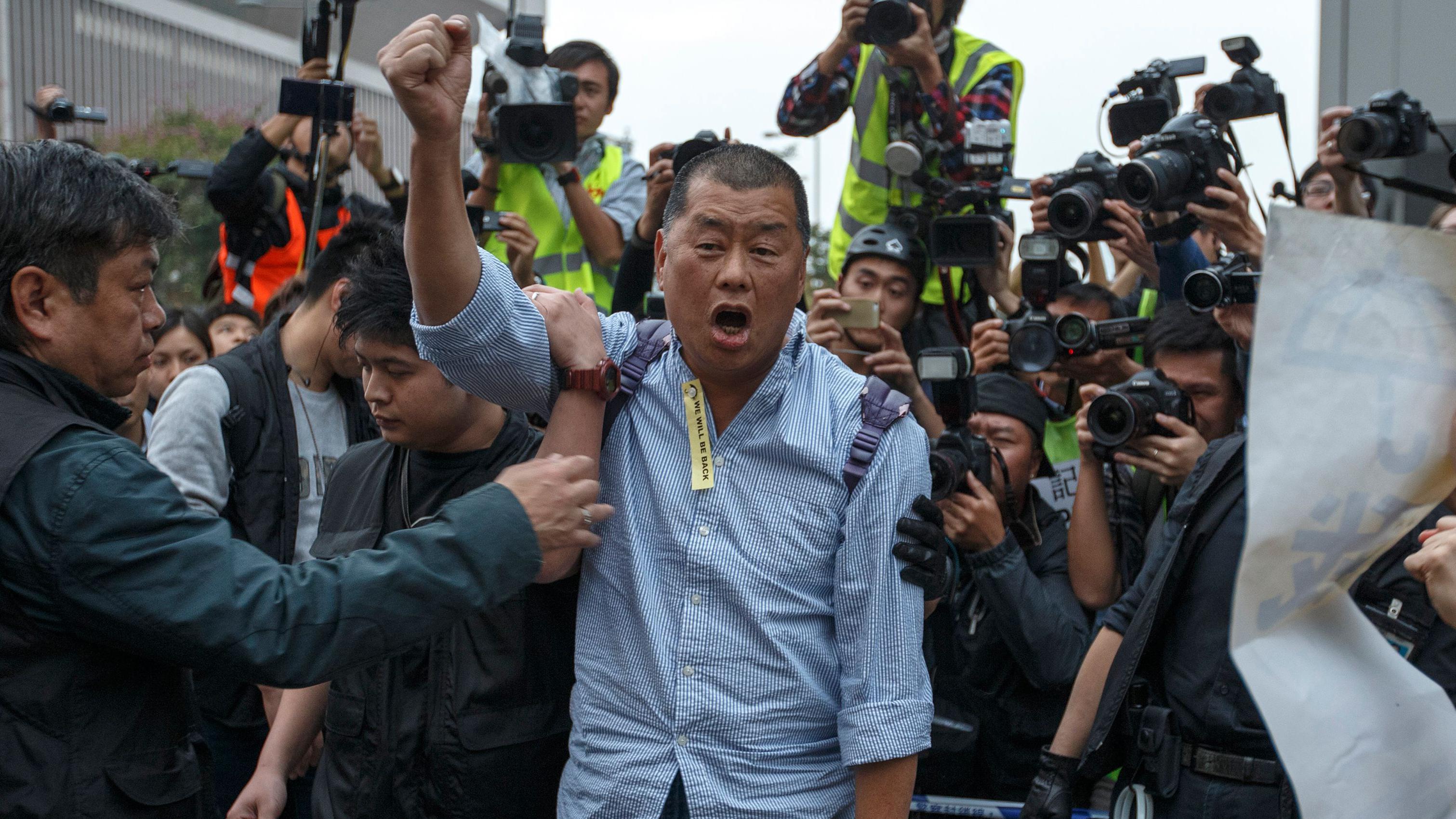 身穿藍色直間條襯衣,右衣襟別上黃色「我們會回來的」小條幅的黎智英在香港金鐘夏愨道上的示威者佔領區內舉著右手叫喊口號,便衣警員準備將其拘捕帶走(11/12/2014)