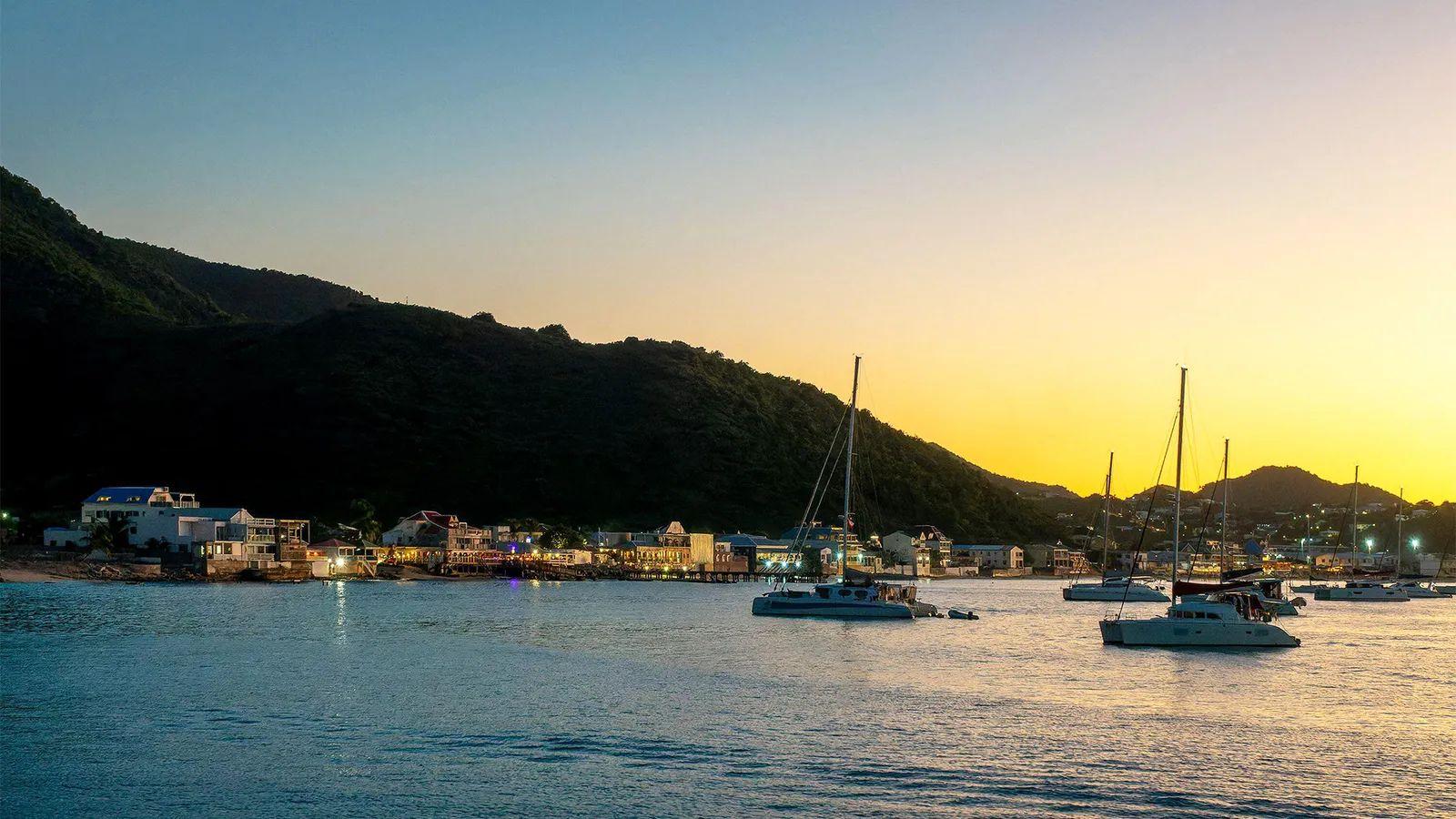 Vista de el mar, buques, la orilla en la distancia con edificaciones y montañas atrás, al atardecer