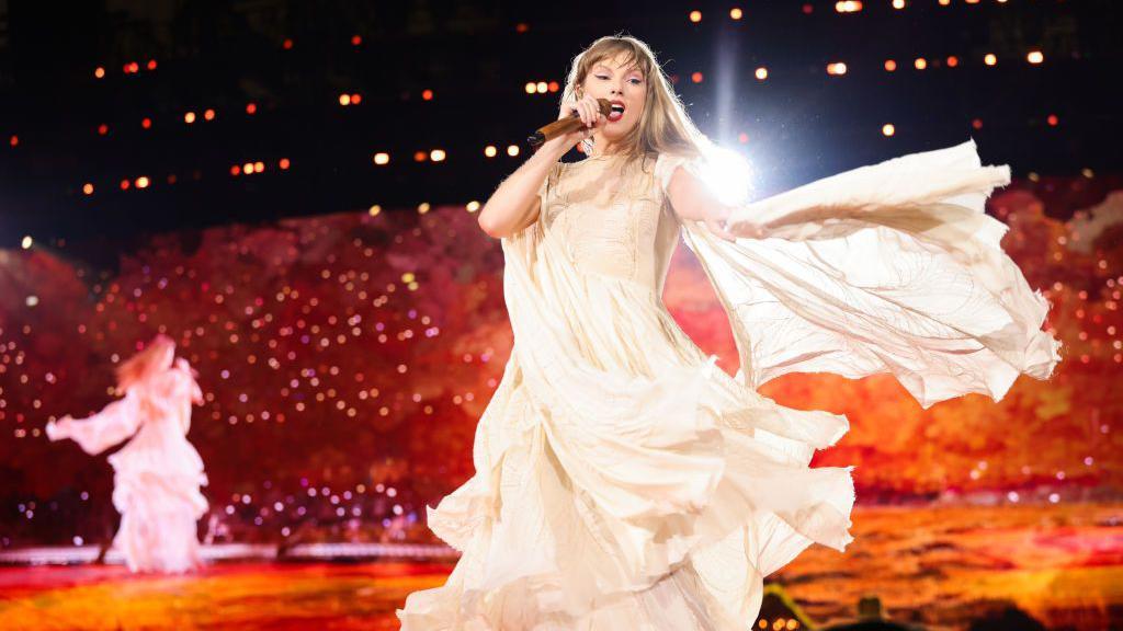 Taylor Swift dança em um vestido de baile na cor creme ornamentado durante sua turnê Eras de 2024