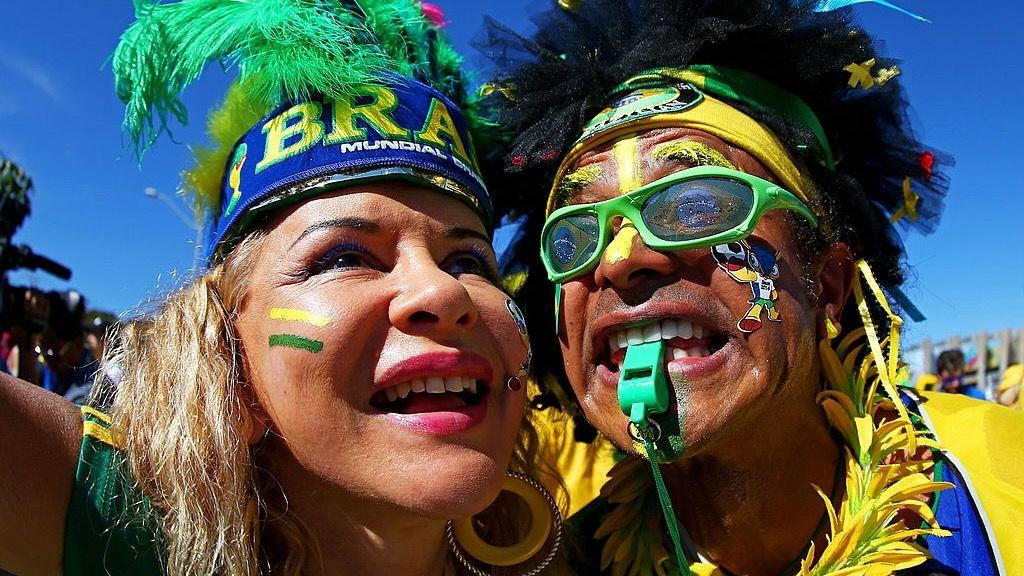 Uma foto em close-up de dois torcedores brasileiros vestindo roupas verdes e amarelas, além de cocares de penas.