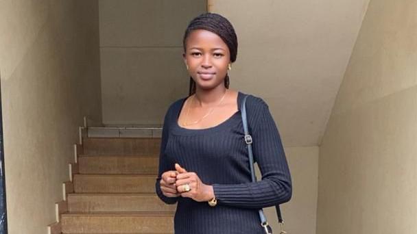 Noella Mukumbi, de p&eacute; em frente a uma escada, com blusa e cal&ccedil;a preta e carregando uma bolsa da mesma cor, sorri para a c&acirc;mera
