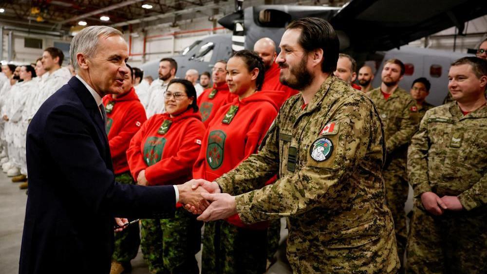 O primeiro-ministro Mark Carney &eacute; visto apertando a m&atilde;o de um membro das For&ccedil;as Armadas Canadenses. Atr&aacute;s deles, fileiras de militares de diferentes unidades est&atilde;o em frente a uma aeronave militar dentro de um hangar.