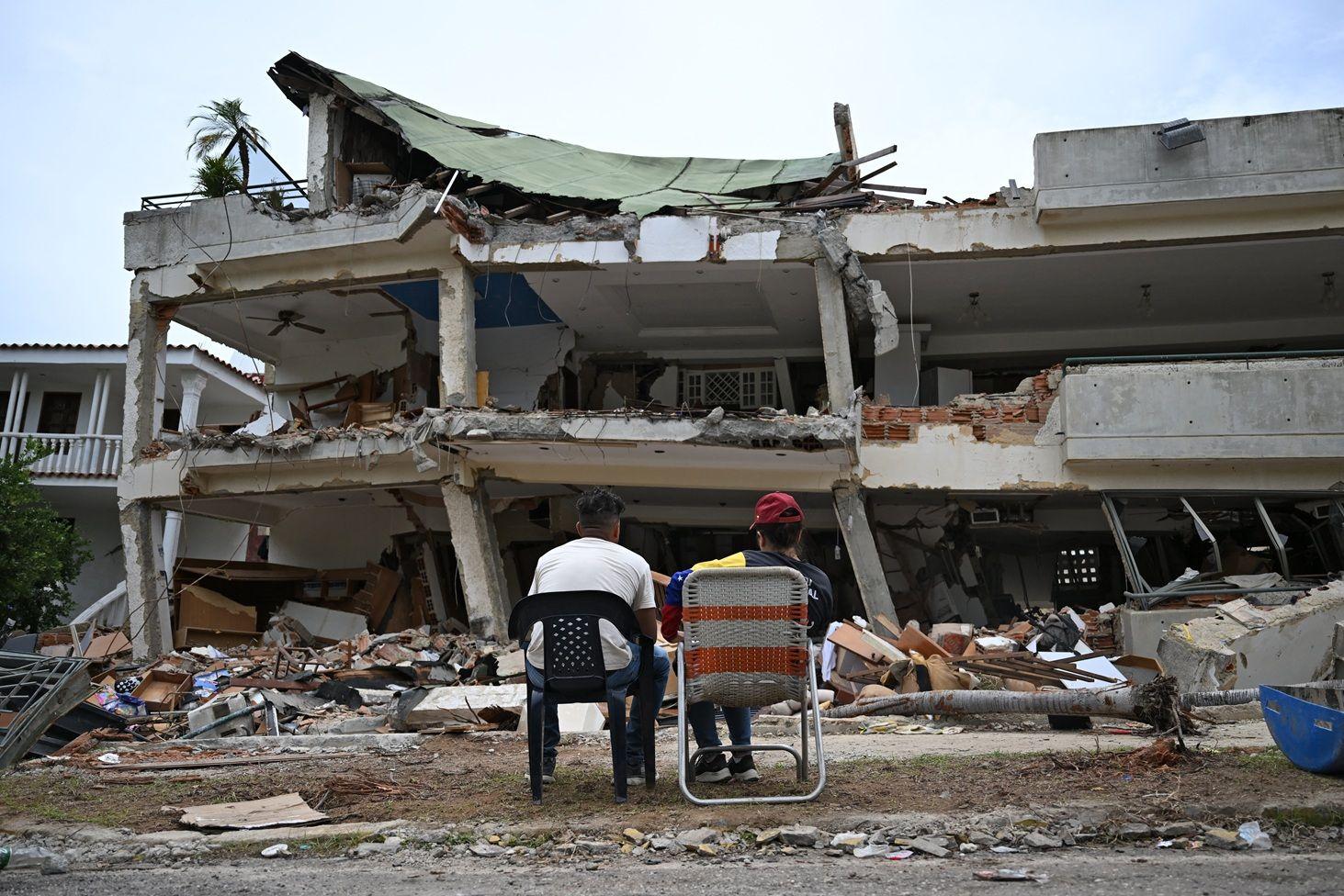 Dos personas están sentadas frente a los edificios de un edificio que quedó destruido
