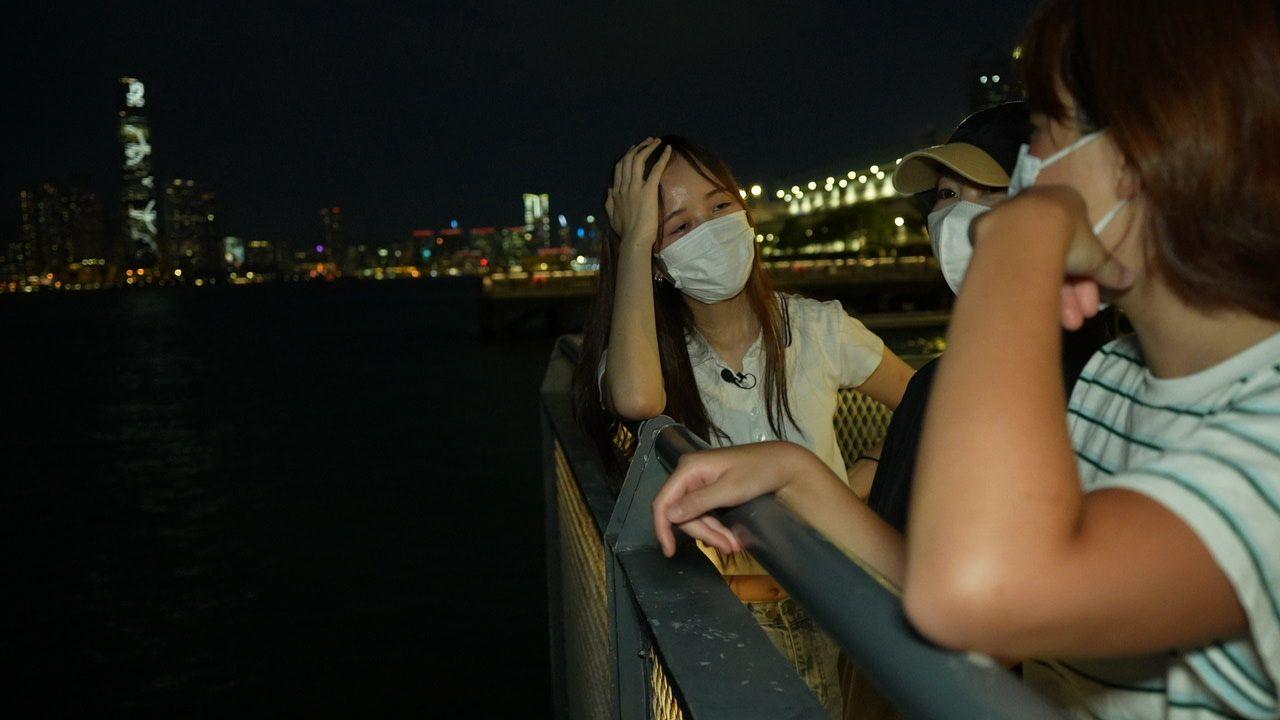 The three women stand at night at the right hand side of the photo talking, leaning on a fence which separates them from the harbour which is pitch black. In the background we see the lights of the city including one tall tower. 