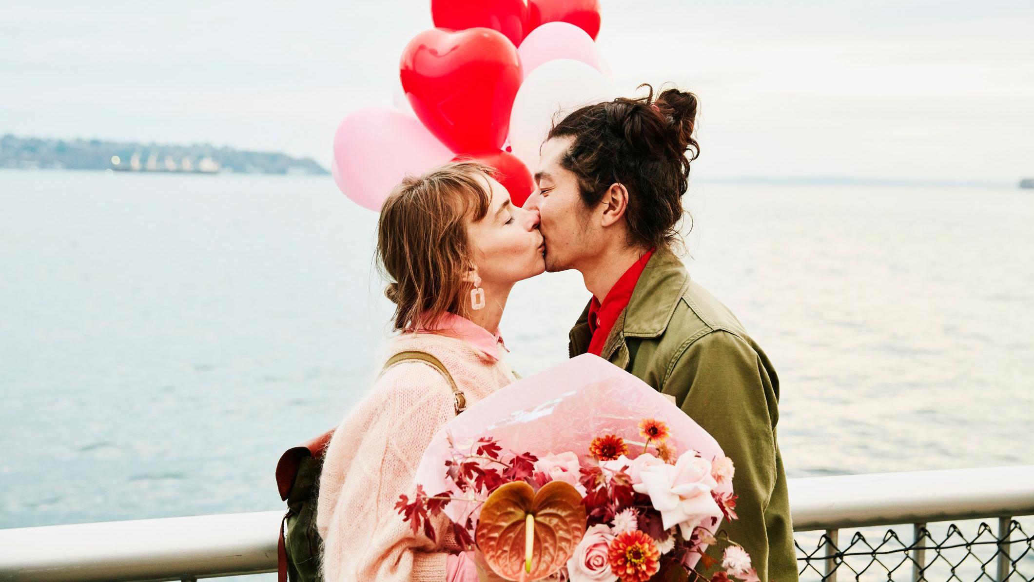 Casal se beijando &agrave; beira-mar, segurando um buqu&ecirc; de flores, com bal&otilde;es vermelhos em formato de cora&ccedil;&atilde;o ao fundo.
