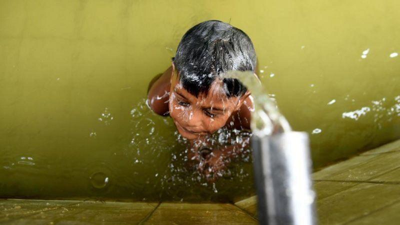 একটি শিশু পানিতে গা ডুবিয়ে আছে, তার মাথায় একটি দল থেকে পানি পড়ছে (ফাইল ছবি)