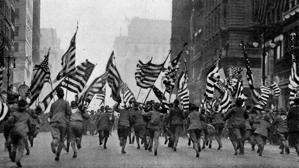 Soldados americanos uniformizados correm por uma rua da cidade de Nova York acenando com as bandeiras de seu pa&iacute;s, em uma fotografia de 1917