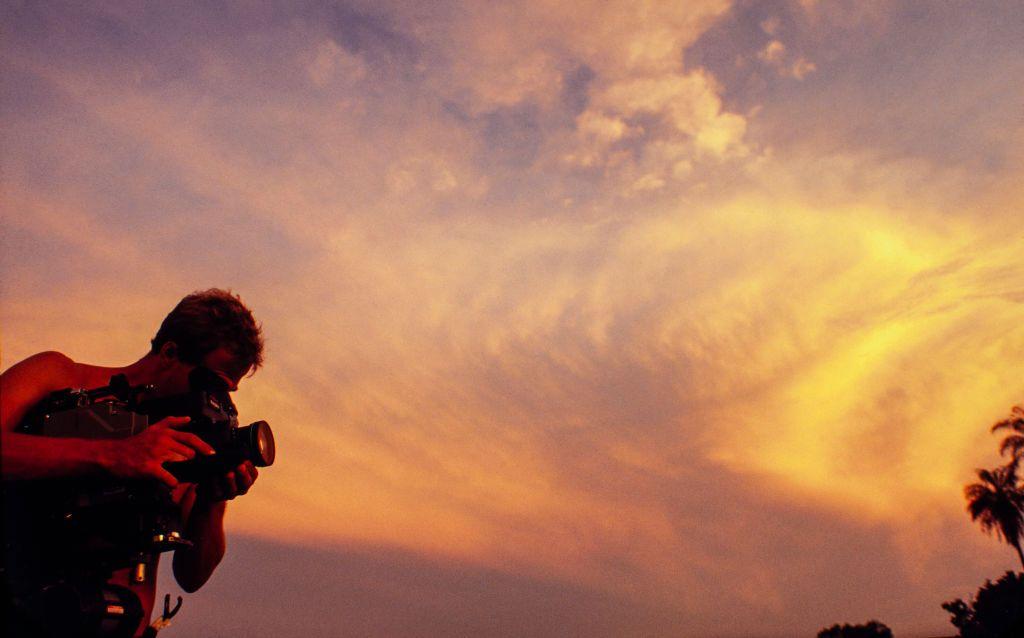 A imagem mostra uma pessoa operando uma c&acirc;mera de v&iacute;deo profissional ao ar livre durante o p&ocirc;r do sol. Em silhueta contra um c&eacute;u alaranjado e repleto de nuvens iluminadas, o cinegrafista aparece concentrado na filmagem, enquanto a luz quente do entardecer domina a composi&ccedil;&atilde;o. Ao fundo, a copa de uma palmeira refor&ccedil;a a atmosfera tropical da cena.