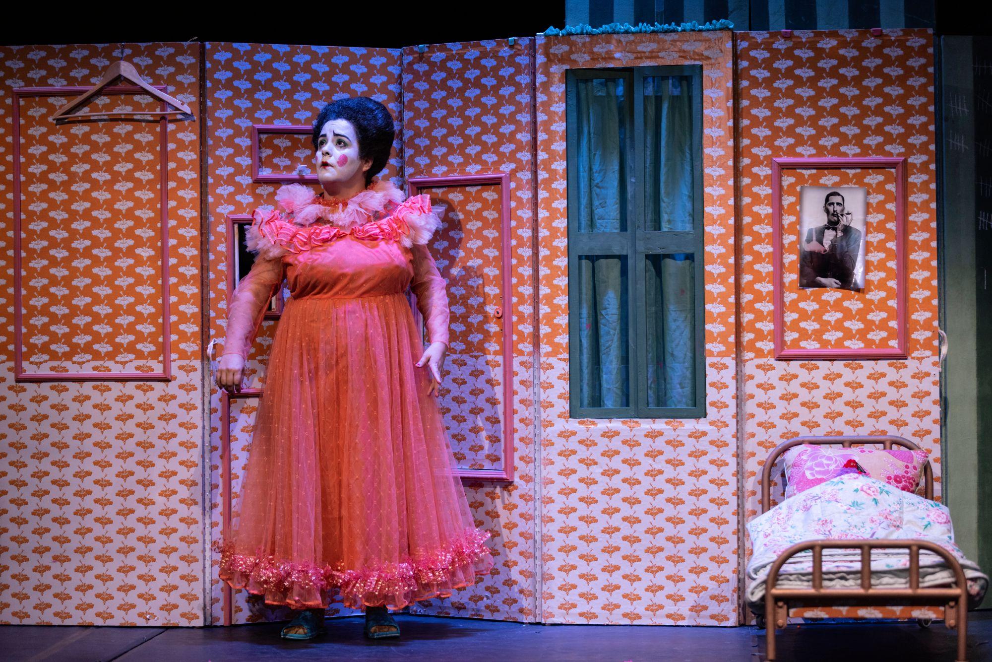 El personaje de Chula, vestida de naranja, frente a una escenografía sencilla que incluye el retrato de un hombre y una cama demasiado pequeña para el personaje