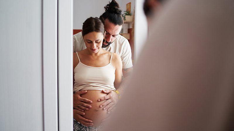 Casal observa no espelho a barriga da mulher, gr&aacute;vida