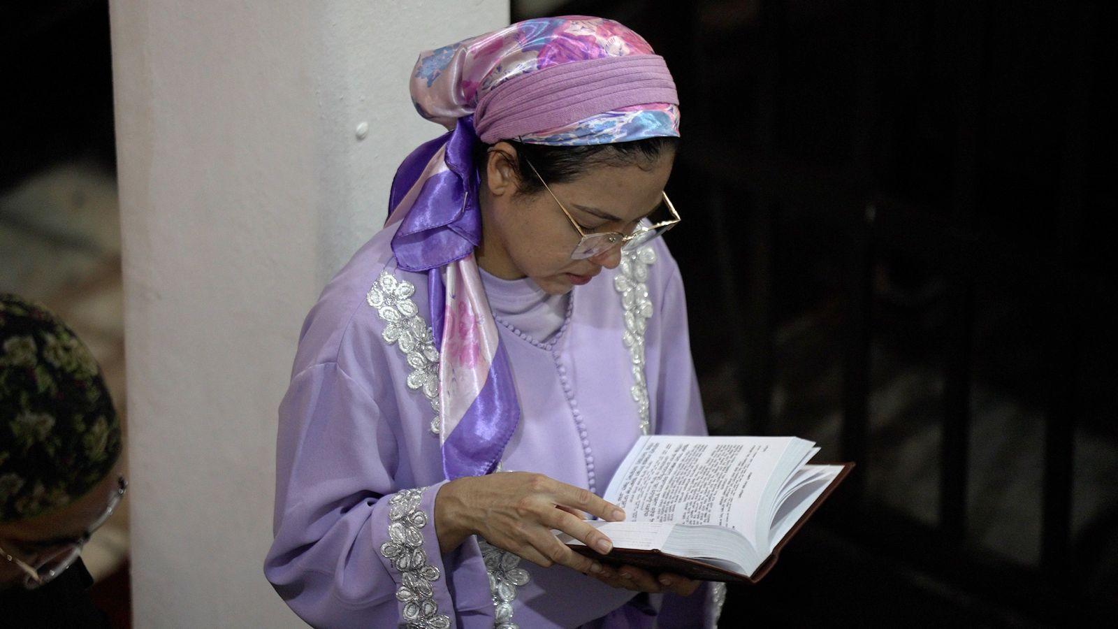 Mulher de óculos com pano rosa na cabeça e roupa lilás lê livro religioso.