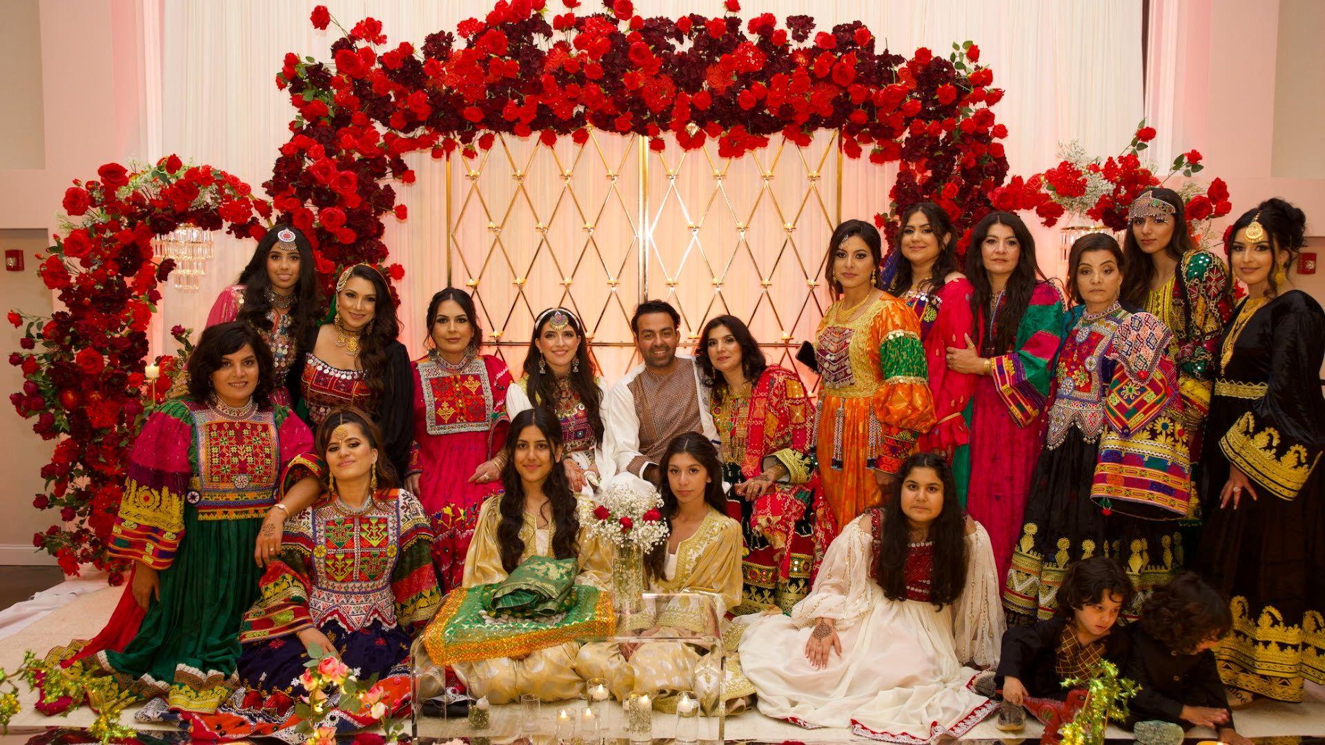 Sammi and Safi posing at their wedding with their guests with a backdrop of red flowers in a banquet hall. Their guests are wearing Afghan attire.