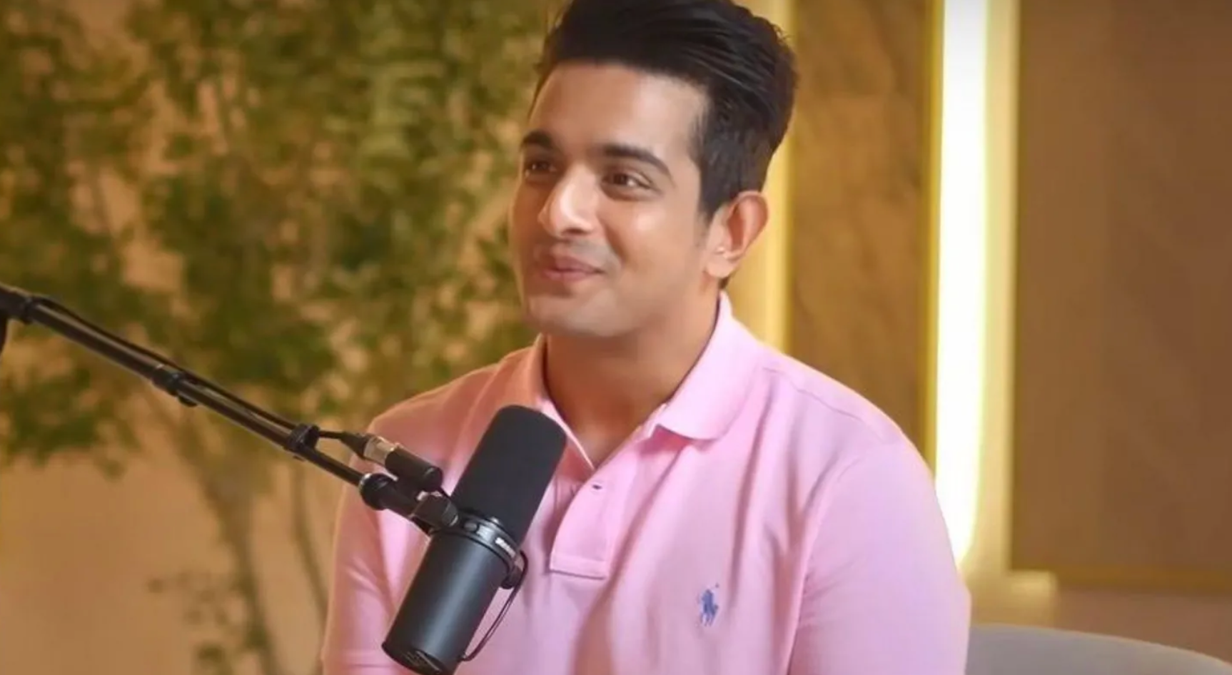 Ranveer Allahbadia during his podcast. He is wearing a light pink polo shirt and is sitting in front of a mic in a warmly lit studio with yellow interiors. 