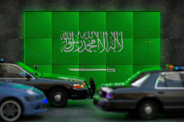 Carros passando em frente a tela com bandeira da Arábia Saudita