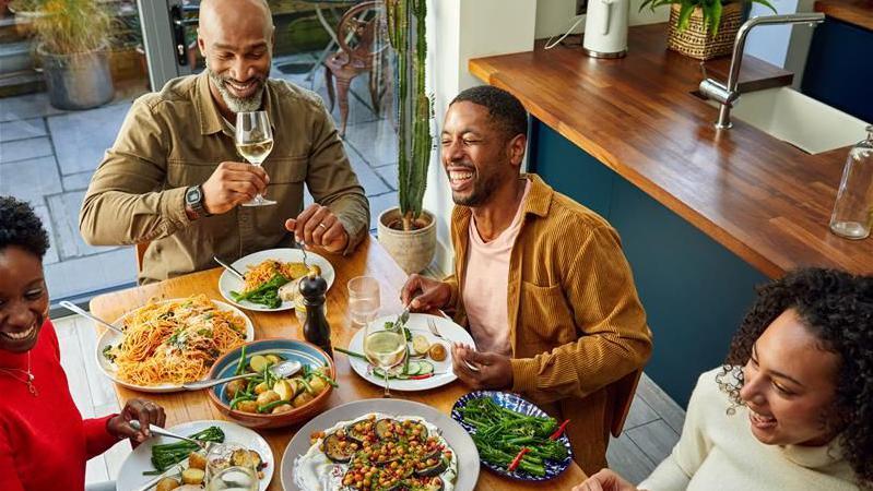 عائلة تبتسم وتضحك وهم يجلسون حول طاولة خشبية يتناولون وجبة طعام. يُقدّم لهم البروكلي والبطاطا والزيتون والسمك والباذنجان. 