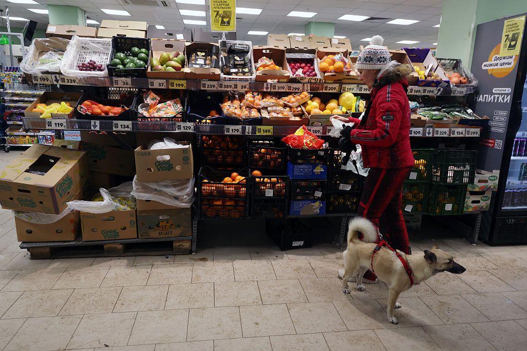 Uma mulher com um casaco de inverno vermelho e um cachorrinho na coleira caminham em dire&ccedil;&atilde;o &agrave; se&ccedil;&atilde;o de frutas do supermercado.
