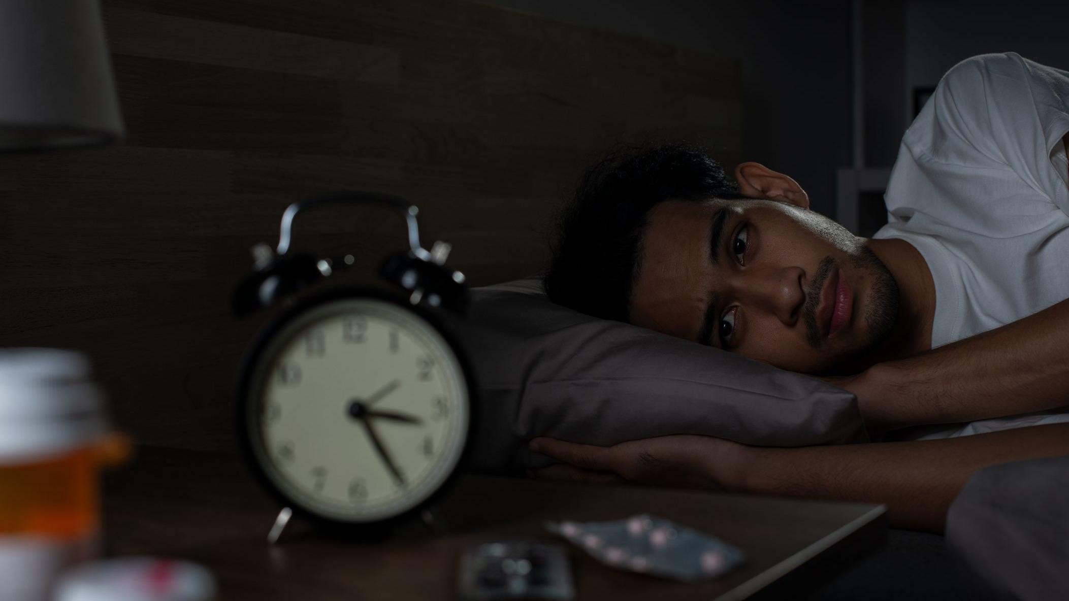 Homem deitado na cama &agrave; noite, acordado, olhando para o despertador que marca 3h25.