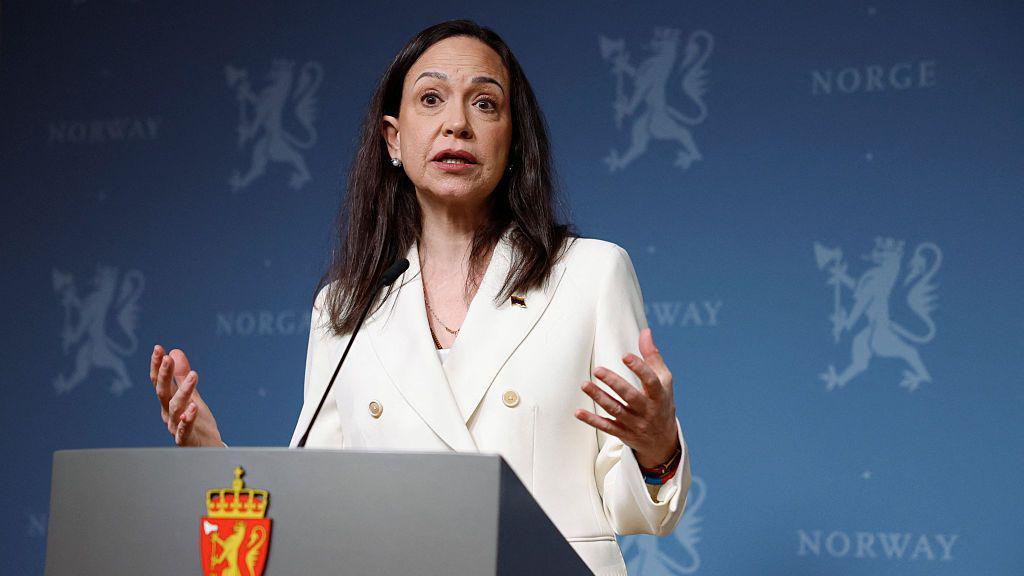 María Corina Machado frente a un podio durante una conferencia de prensa en noruega. Lleva el cabello negro hasta los hombros. Tiene una expresión seria mientras habla. Tiene las manos extendidas hacia los lados. Viste un chaleco blanco. 