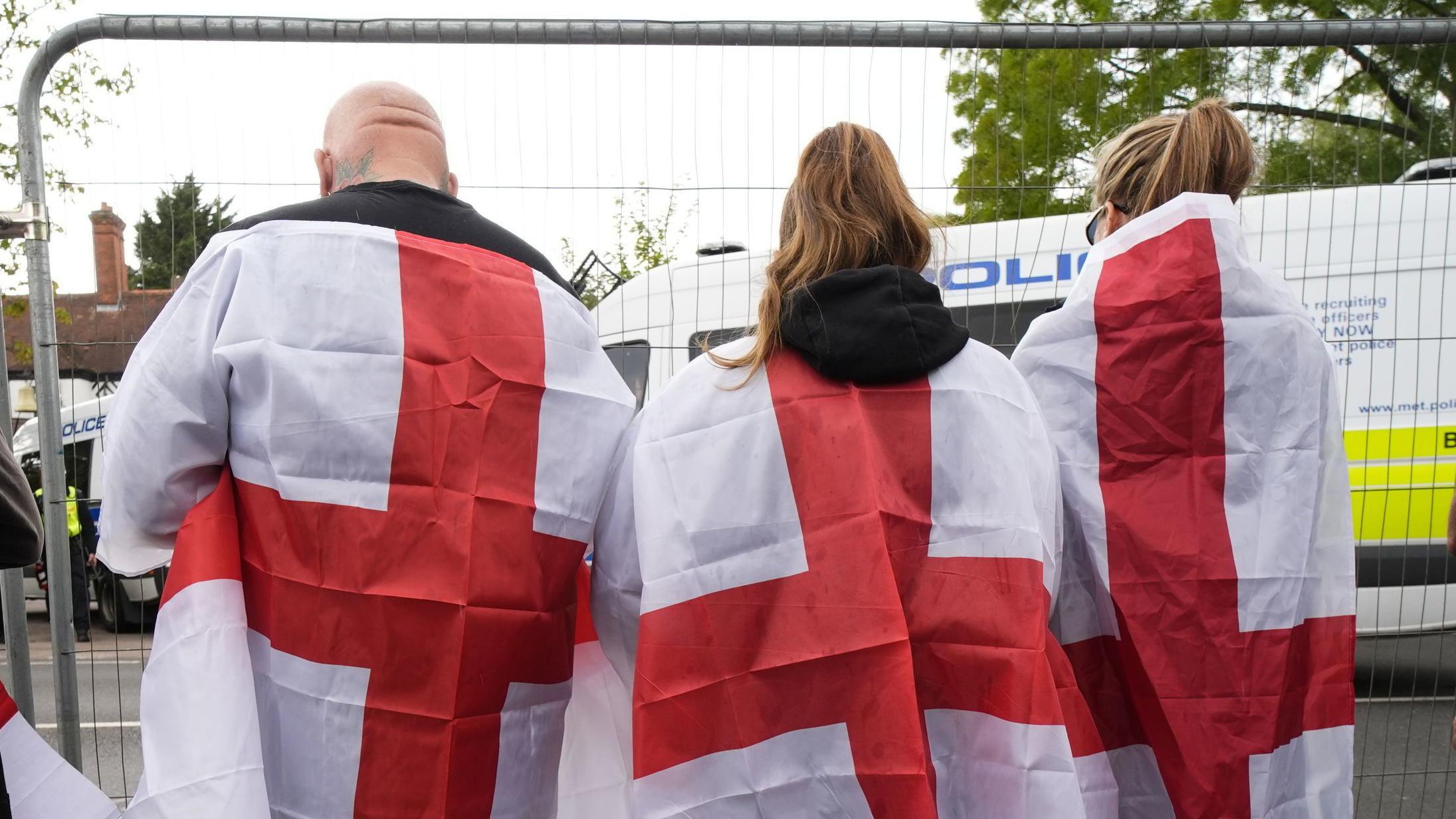 Cuatro personas, tres de las cuales llevan la bandera de San Jorge sobre sus hombros, de pie detrás de una valla metálica; frente a ellas se encuentra el Bell Hotel, en Epping, Essex, sureste de Inglaterra, que está bloqueado por furgones policiales.