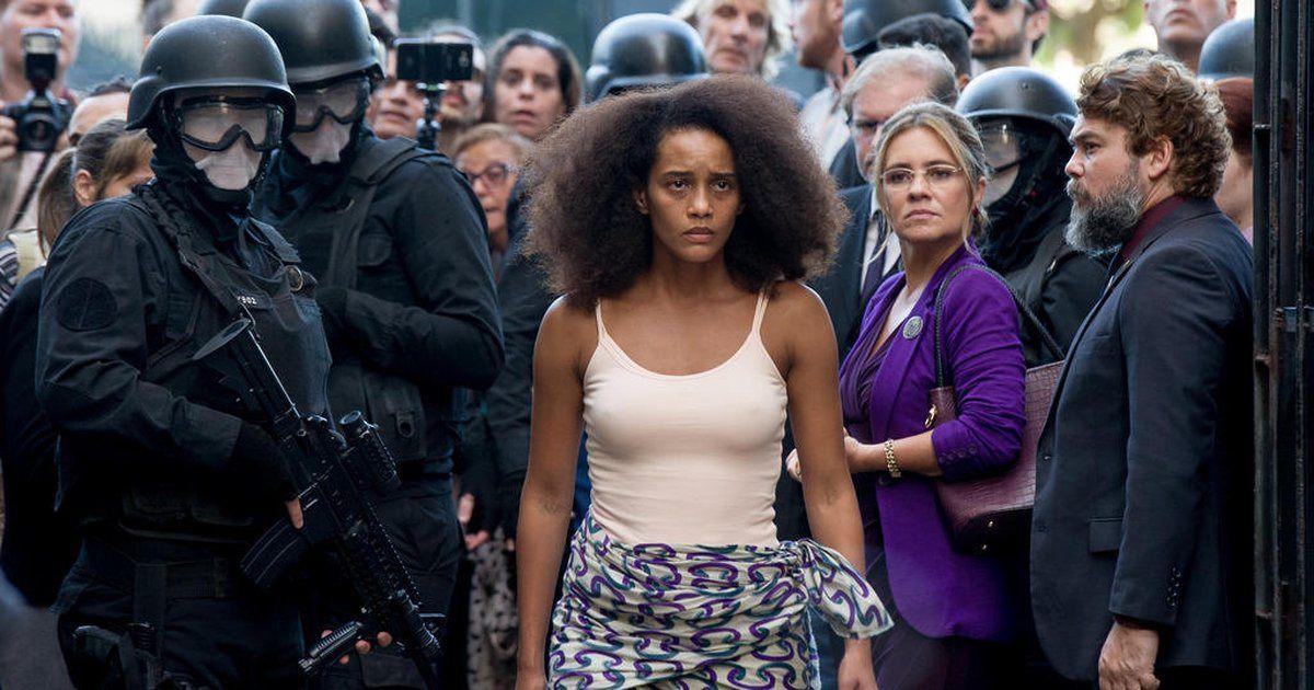 Uma mulher negra, a atriz Ta&iacute;s Ara&uacute;jo, caminha entre policiais armados, enquanto uma multid&atilde;o observa a cena ao fundo, criando um clima de tens&atilde;o e confronto em cena do filme Medida Provis&oacute;ria.