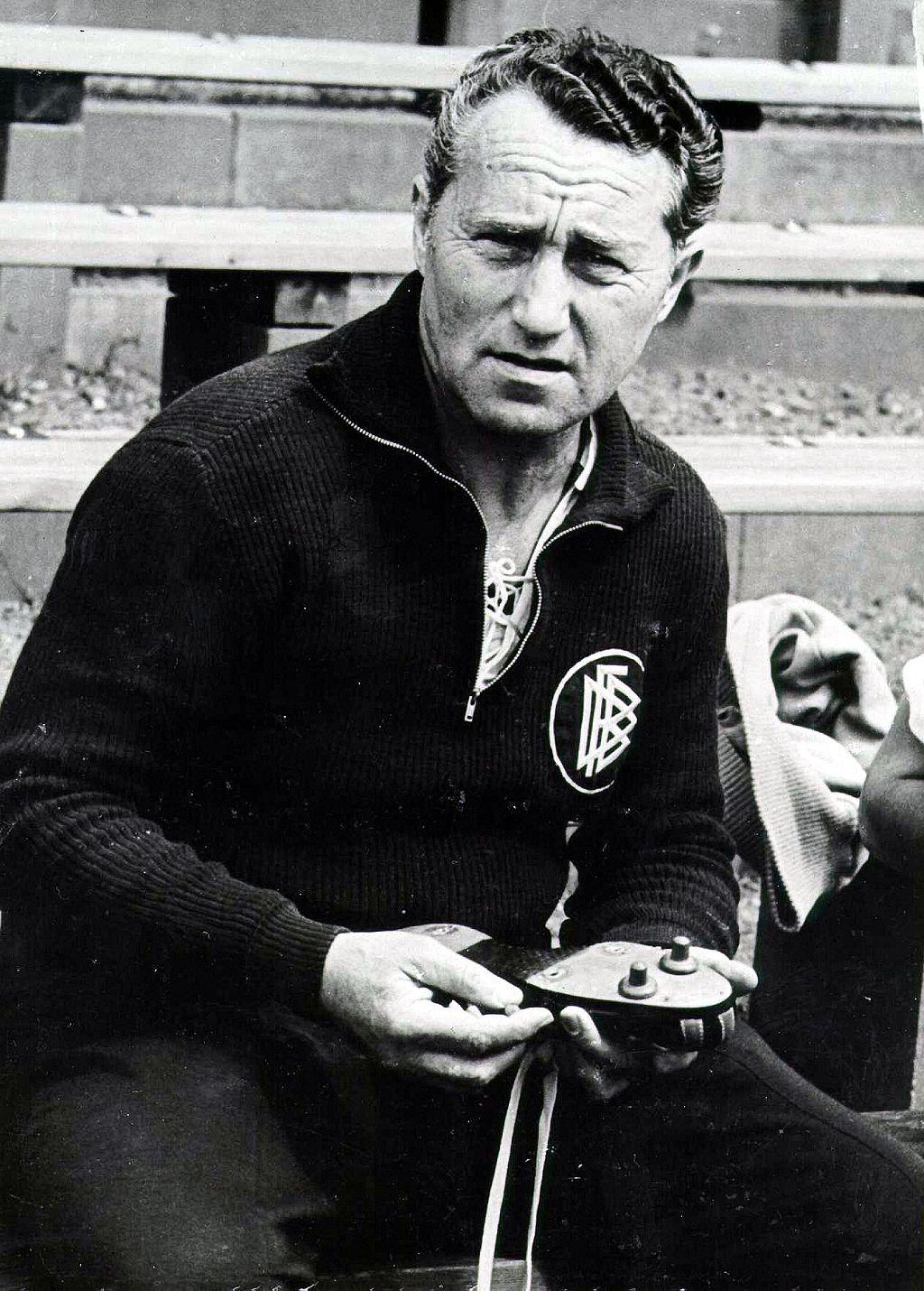 Fotografía en blanco y negro de Adolf Dassler, fundador de la empresa alemana de ropa deportiva Adidas, con ropa oscura y la insignia de la Federación Alemana de Fútbol (DFB) a un lado en el pecho, sosteniendo zapatillas de fútbol con dos tacos y huecos para fijar más. 