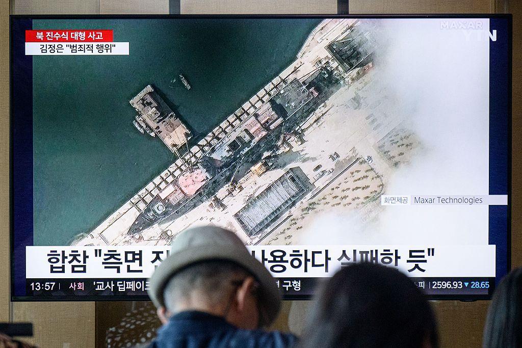En una fotografía tomada en Seúl el 22 de mayo de 2025, la gente se sienta cerca de un televisor que muestra imágenes de noticias con una imagen satelital cortesía de Maxar Technologies que muestra una vista general de un nuevo buque de guerra norcoreano en el puerto antes de su botadura en Chongjin.