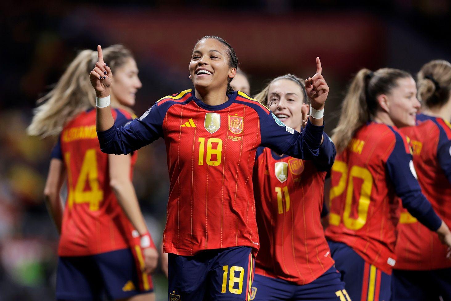 Vicky López, vestida con el uniforme de la selección española, sonríe mientras sus dedos índices apuntan hacia el cielo. En el fondo se ven otras futbolistas españolas

