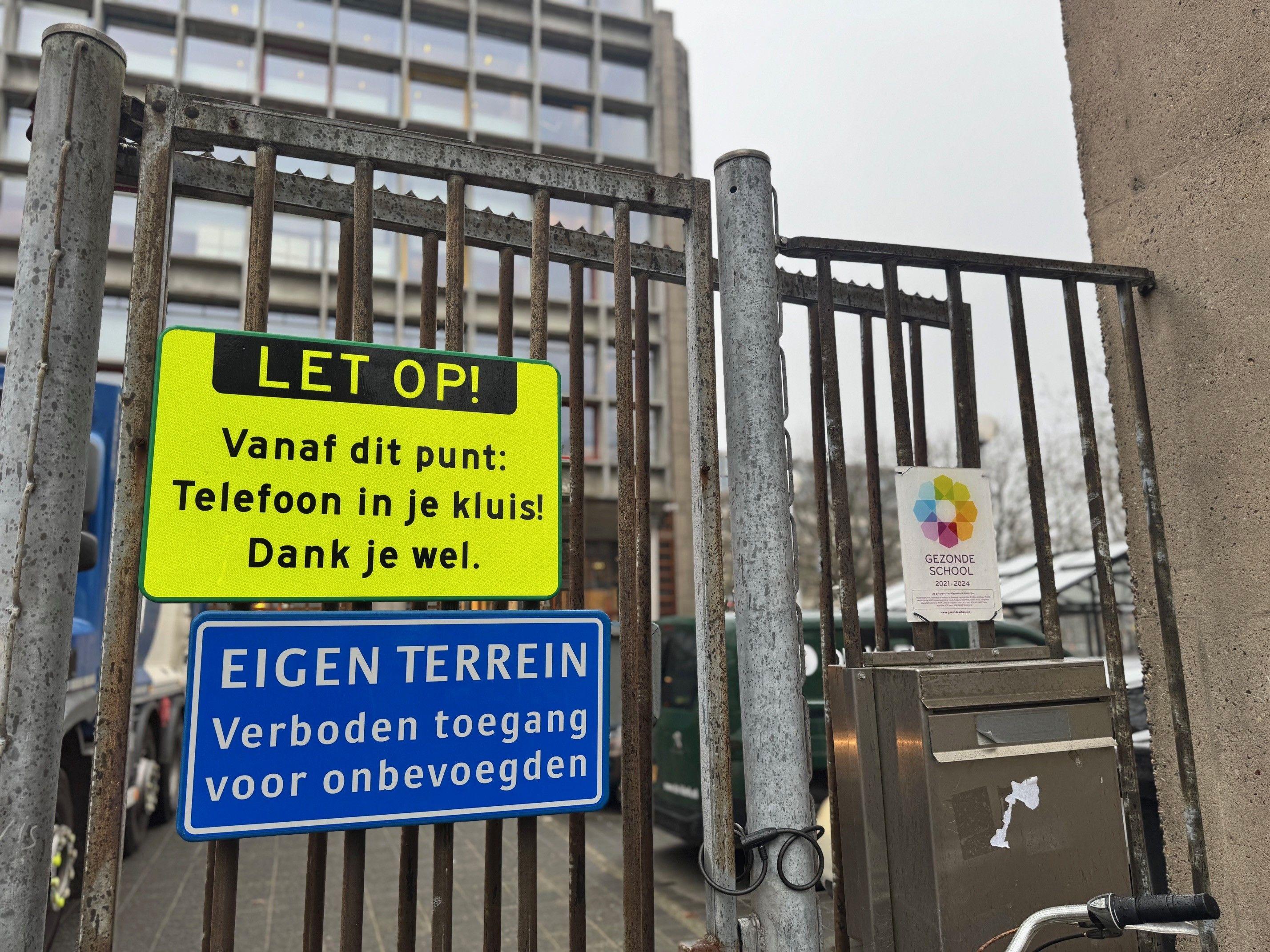 Una valla metálica rodea una escuela con un cartel amarillo brillante que indica a los alumnos que dejen sus teléfonos en sus taquillas.