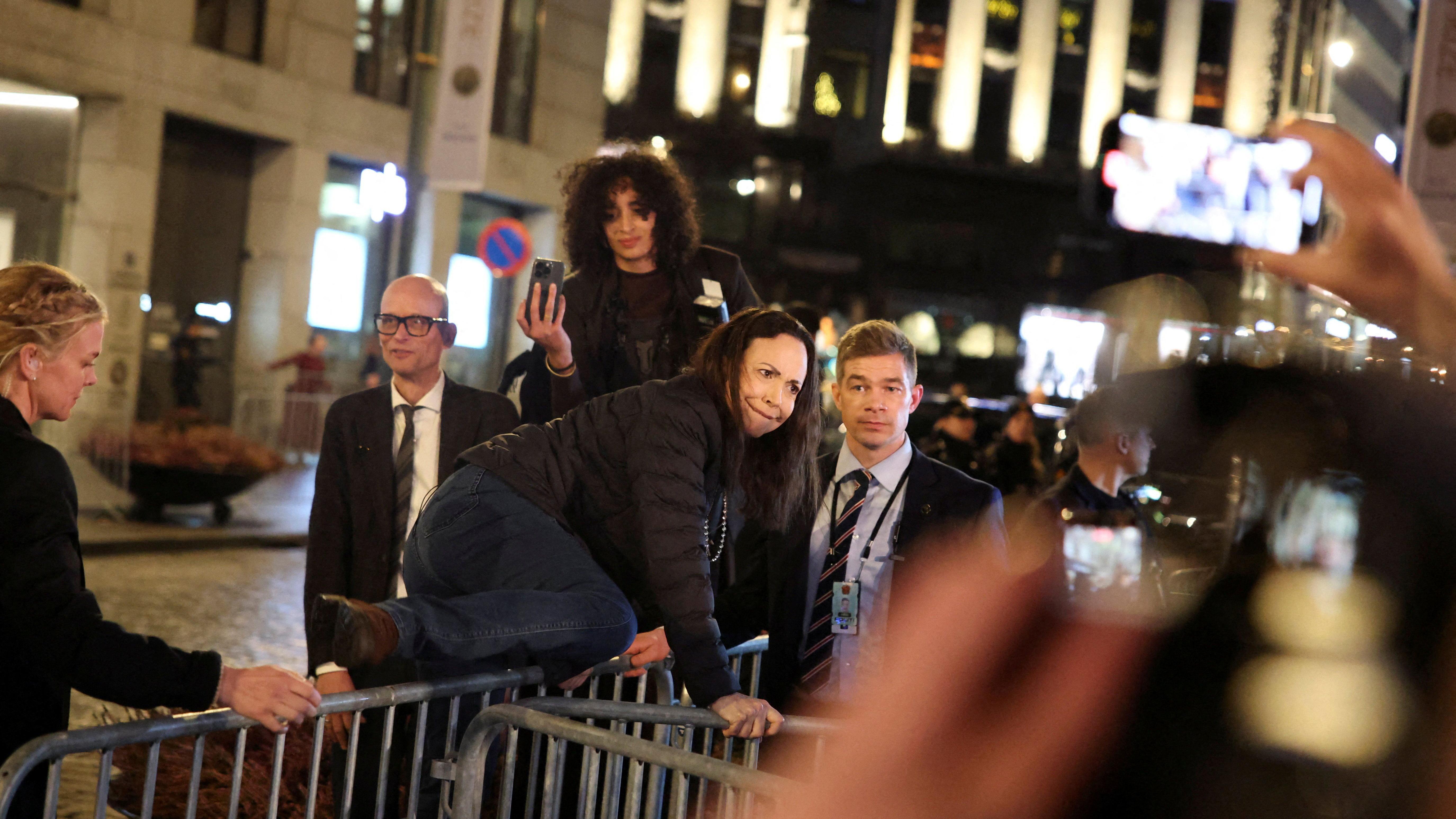 María Corina Machado salta as grades de segurança diante do Grand Hotel, em Oslo, para cumprimentar apoiadores que aplaudem, enquanto seguranças observam