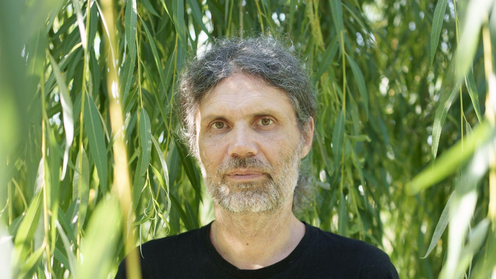 Muškarac sa dužom kosom gleda u kameru, iza njega je zelenilo, ima sedu kosu i bradu