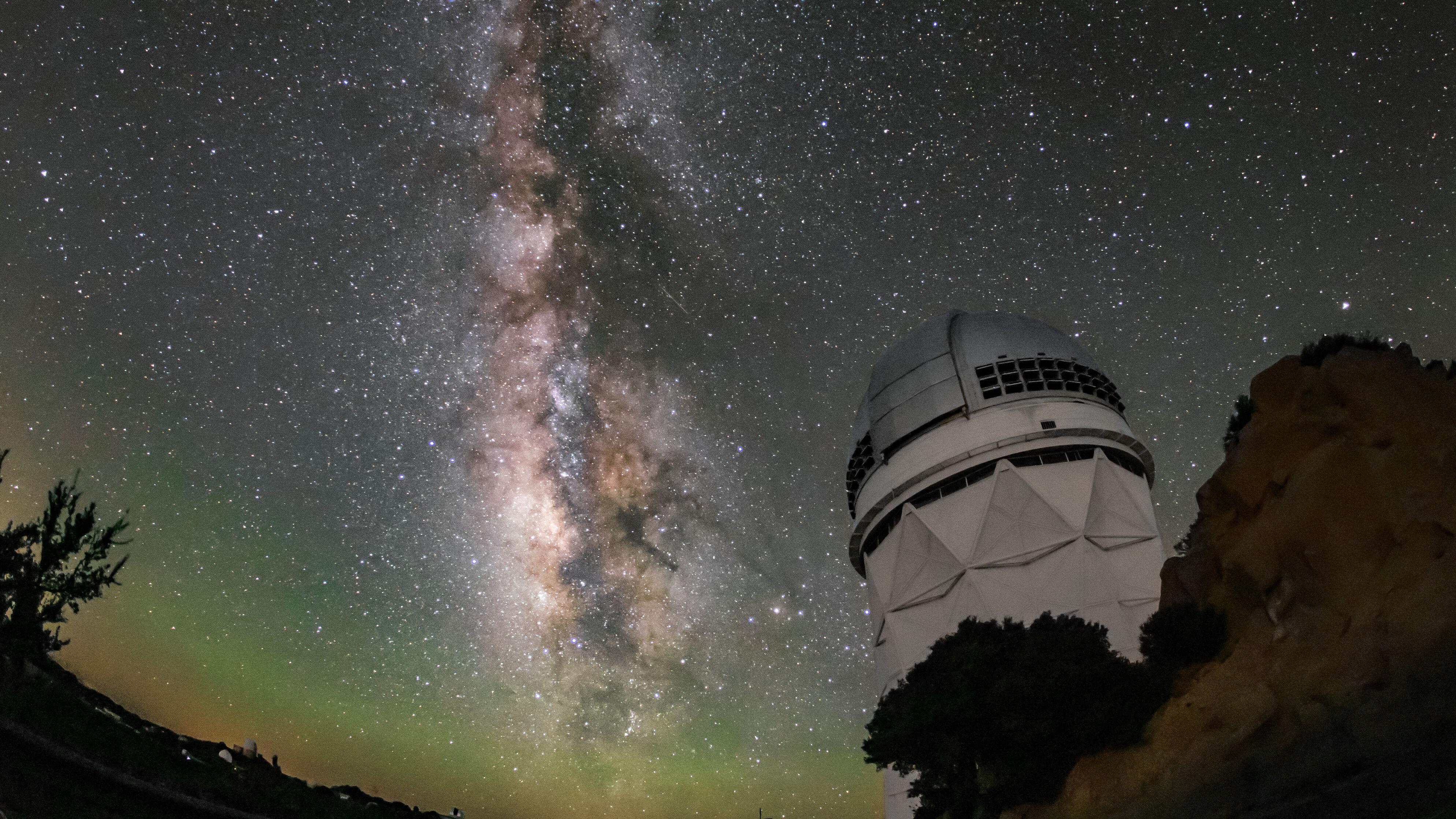 A tall, white-domed telescope stands on a rocky hillside under a perfectly clear night sky.Next to it, the Milky Way appears as a bright, vertical river of light, mottled with dense star clouds and dark dust lanes, stretching from the bottom to the top of the picture.The foreground is mostly in shadow, with a few dark trees silhouetted against a faint greenish glow near the horizon.